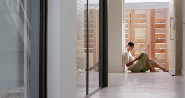 A man is sitting alone on the floor in a modern hallway near an open patio door. The scene showcases a relaxed and casual atmosphere with modern minimalist design elements. Potential uses include lifestyle blogs, articles about relaxation or living spaces, and interior design inspiration.