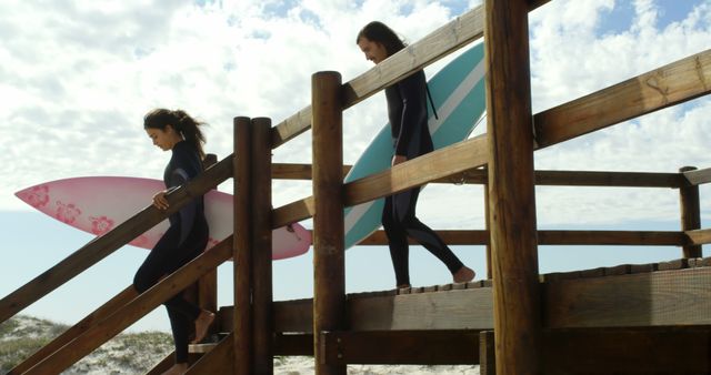 Two women wearing wetsuits are walking down a wooden staircase towards the beach, carrying their surfboards. The sky is partly cloudy and the atmosphere signals a perfect day for surfing. This vibrant and lively scene can be used in promotions for surf schools, outdoor adventure tours, summer vacation destinations, and lifestyle blogs.