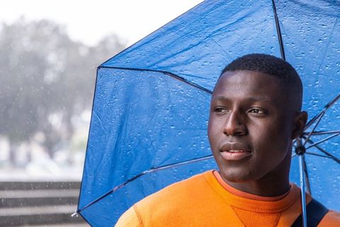 Young man under blue umbrella in rain wearing orange sweater, urban contemplative portrait