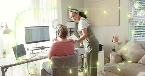 Two women collaborating and reviewing charts at home workspace with monitor and laptop