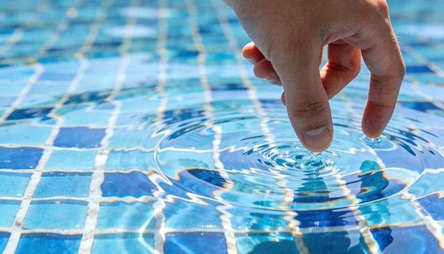 Index finger creating concentric ripples over blue mosaic pool tiles, close-up water texture