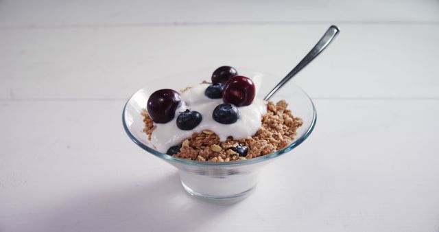 A bowl of yogurt with granola and fresh berries is served on a white surface, with copy space. Ideal for a healthy breakfast or snack, the dish offers a nutritious combination of dairy, whole grains, and fruit.