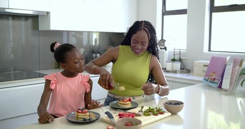 African american mother pouring syrup on pancakes with smiling daughter in sunlit kitchen