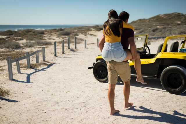 Couple enjoying a sunny day at the beach, with one person giving the other a piggyback ride towards a yellow beach buggy. Ideal for use in travel and tourism promotions, summer holiday advertisements, romantic getaway brochures, and lifestyle blogs focusing on adventure and outdoor activities.