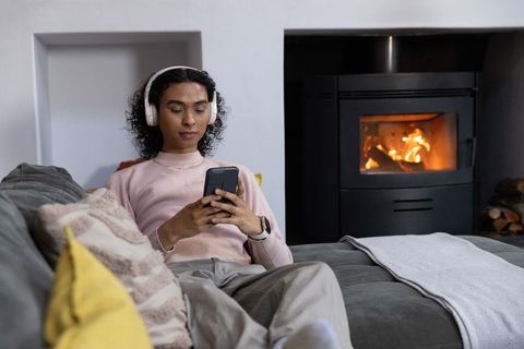 Nonbinary person relaxing on gray sofa with headphones and smartphone by cozy wood stove