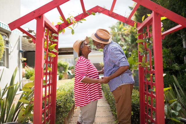 Senior couple standing in a garden, holding hands and smiling at each other. They are surrounded by greenery and a red garden arch. This image can be used for themes related to love, relationships, retirement, and outdoor activities for seniors.