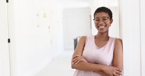 Confident african woman smiling while leaning in bright minimalist hallway with glasses and pink top
