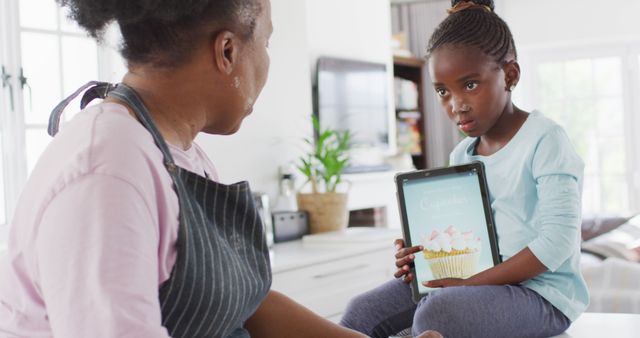 Elderly African American woman showing young girl a baking recipe using a tablet. Two generations are bonding over cooking in a bright, modern kitchen. Ideal for topics on family, education, baking, technology in the kitchen, and intergenerational relationships.