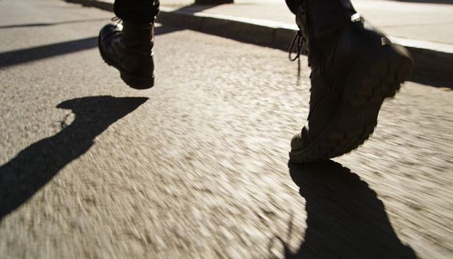 Striding in heavy black lace-up combat boots on sunlit urban asphalt with dramatic shadow