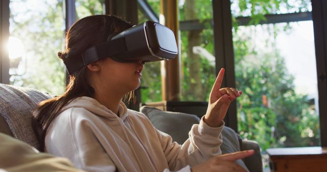 Young woman wearing a virtual reality headset, exploring an immersive experience in a cozy living room. Ideal for use in technology blogs, articles about modern lifestyle, virtual reality businesses, and advertisements for VR products.