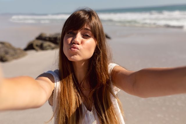 Young woman pouting while taking a selfie on a sunny beach. Ideal for use in travel blogs, vacation advertisements, lifestyle magazines, and social media content promoting summer fun and beach activities.