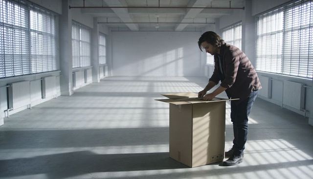 Man unpacking cardboard box in sunlit industrial loft with blinds and grid shadows