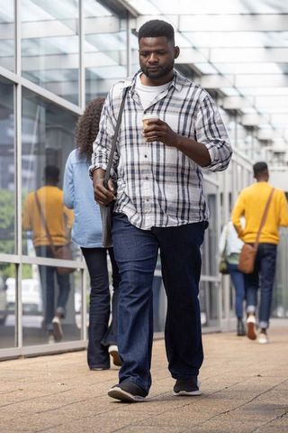 African commuter walking with to-go coffee and messenger bag along glass-covered walkway