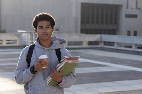 Sunlit african american teen student holding coffee and notebooks on urban campus rooftop