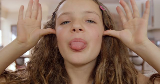 Young girl with long hair making silly face by sticking out tongue and placing hands by cheeks. Fun and whimsical moment indoors. Ideal for use in children's activities, lifestyle blogs, and playful advertisement campaigns targeting families or creating a light-hearted atmosphere.