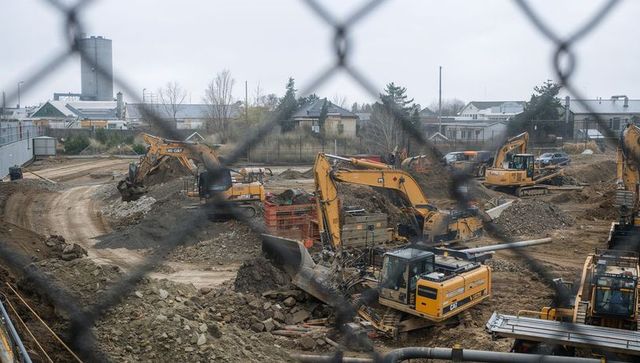 Excavators digging urban construction site through chainlink fence, crews working