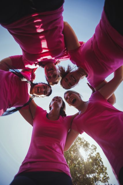 Group of women forming a huddle during a boot camp on a sunny day. They are wearing pink shirts, symbolizing unity and teamwork. This image can be used for promoting fitness programs, team-building activities, women's empowerment events, and outdoor exercise campaigns.