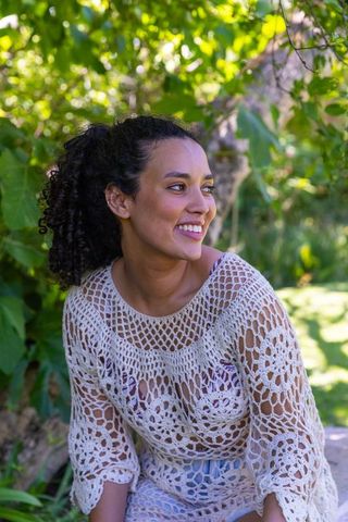African-american woman smiling while sitting in garden wearing cream crochet top and bikini
