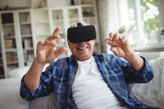 Senior man wearing VR headset, engaging with virtual environment in living room. Ideal for illustrating technology use among elderly, modern lifestyle, and digital innovation. Suitable for articles on virtual reality, senior engagement with technology, and home entertainment.