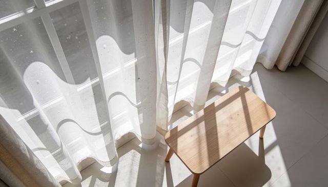 Sunlit sheer curtain casting wavy shadows over minimal wooden stool in bright morning