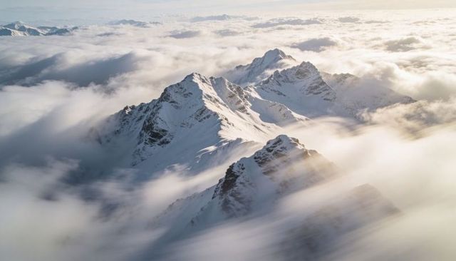 Aerial snowy alpine ridge piercing sunlit sea of clouds with jagged summits and frost