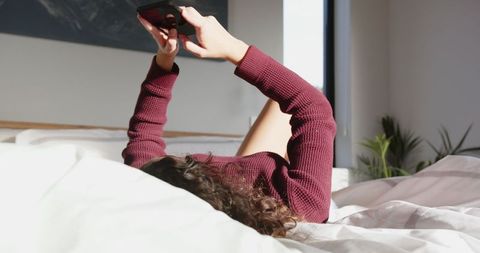 Young Woman Relaxing on Bed with Smartphone in Sunlit Bedroom