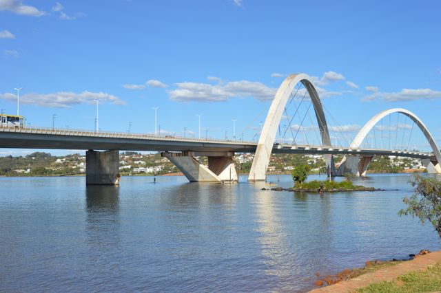 This image features the iconic JK Bridge in Brasília, Brazil, extending over the tranquil Paranoá Lake under a clear sky. The modern architectural design of the bridge with its striking arches is emphasized. This image is ideal for use in travel blogs, architectural publications, and cityscape-related content.