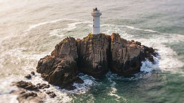 Solitary white lighthouse standing on jagged granite islet with waves crashing at sunset