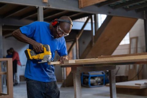 Black craftsman cutting wood plank with cordless jigsaw in carpentry workshop