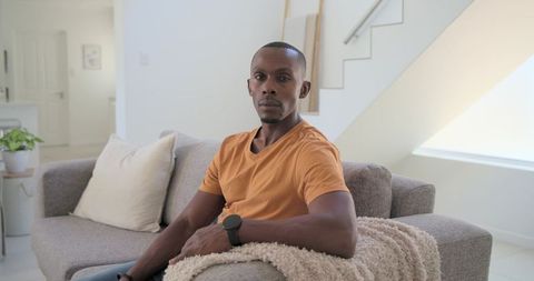 Thoughtful man wearing orange t-shirt sitting on modern sofa in minimalist room