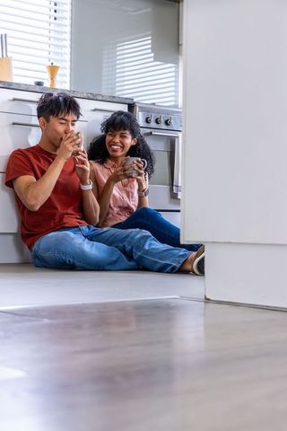 Sunlit couple relaxing on kitchen floor with mugs, asian man and african american woman