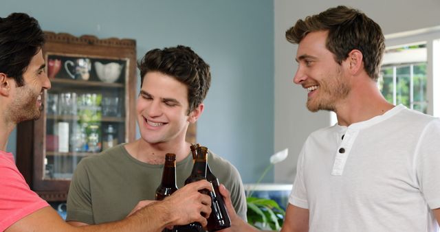 Three smiling men, holding bottles, toasting and enjoying a drink together in a relaxed indoor environment. Suitable for use in themes related to friendship, social gatherings, celebrations, leisure activities, and casual home settings. Perfect for promoting social events, beverage brands, and lifestyle content.