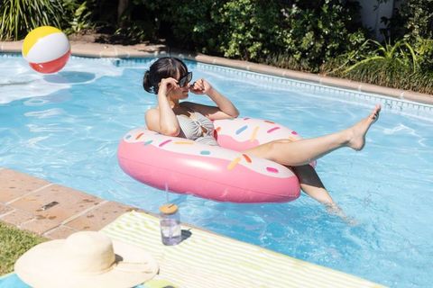 Asian woman relaxing on pink donut float in backyard pool wearing sunglasses and bikini