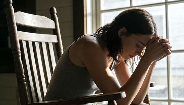 Young woman leaning forward clasping hands in wooden rocking chair by sunlit window