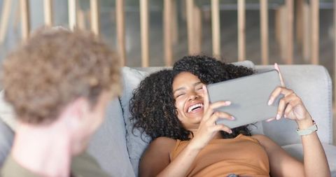 Laughing woman reclining on sofa holding tablet, enjoying relaxed cozy living room moment