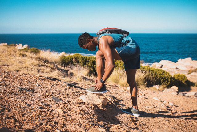 African American man hiking on a coastal trail, tying his shoe. Ideal for promoting outdoor fitness, healthy lifestyle, adventure tourism, and athletic training. Perfect for use in fitness blogs, travel websites, and health magazines.