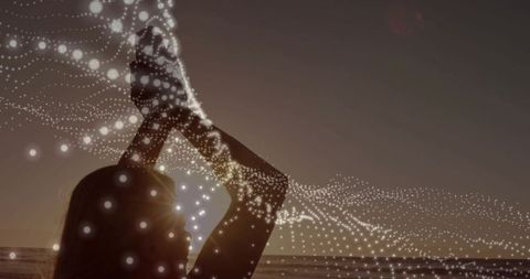 Silhouetting woman raising hands at sunset with glowing dotted data wave over ocean horizon