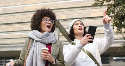 Friends Exploring Urban Sights Pointing Up While Checking Phone and Holding Coffee To-Go