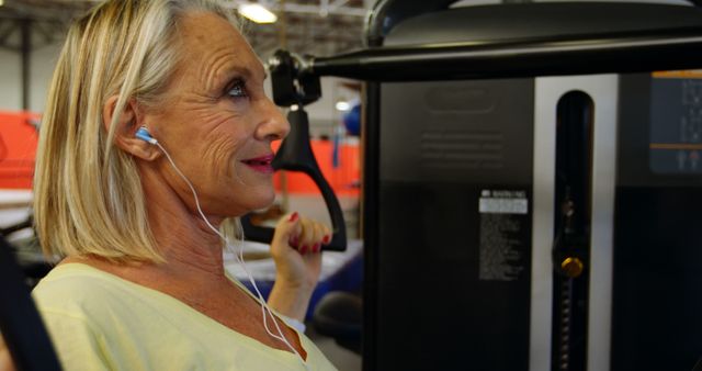 Senior woman with white hair using weight machine in gym while wearing headphones. She is smiling, suggesting enjoyment and commitment to healthy, active lifestyle. Suitable for articles on senior fitness, active aging, exercise routines, healthcare advertisements, and motivational content for mature audiences.