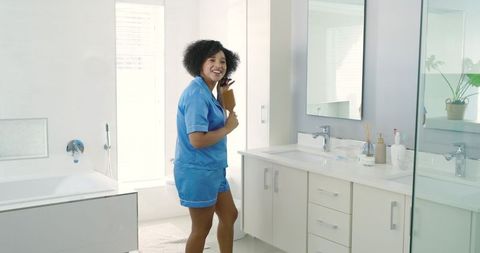 African American woman brushing hair and smiling at bright modern bathroom vanity