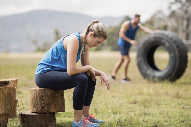 Woman taking a break during an intense outdoor boot camp session. She is sitting on a tree trunk, looking exhausted. In the background, a man is flipping a large tire, indicating a rigorous workout. Ideal for use in fitness blogs, workout motivation content, and advertisements for outdoor training programs.