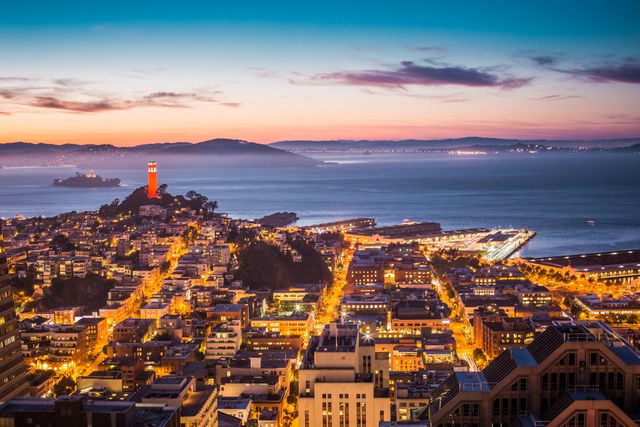 San Francisco cityscape showcasing Coit Tower at sunset, with illuminated streets and the surrounding bay. Ideal for use in travel brochures, city guides, websites about San Francisco, or articles related to urban living and west coast tourism.