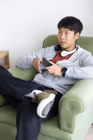 Young asian man lounging in green armchair using smartphone wearing headphones