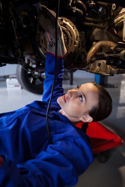 Female mechanic wearing blue coveralls repairing a car from underneath in a garage workshop. Useful for illustrating themes such as women in trades, automotive repair, mechanical engineering, and gender diversity in the workplace.