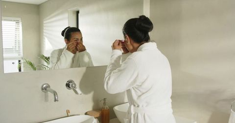 Asian woman standing at bathroom vanity in white bathrobe adjusting contact lenses