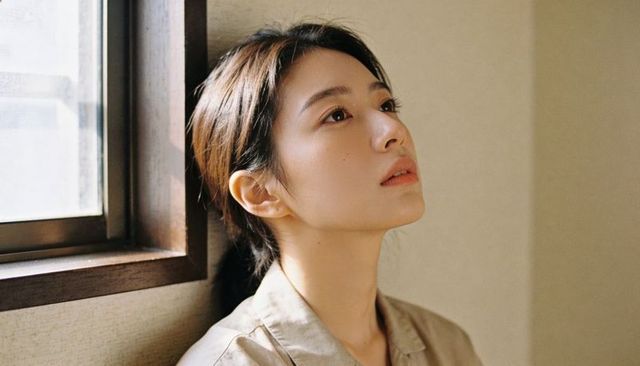 Gazing woman tilting head by sunlit window wearing light beige shirt contemplative portrait