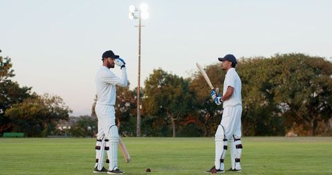 Two diverse cricketers preparing to bat at dusk under glowing floodlights on green field