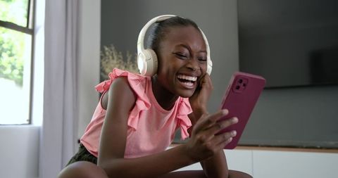 Laughing african girl listening to music on headphones holding smartphone in living room