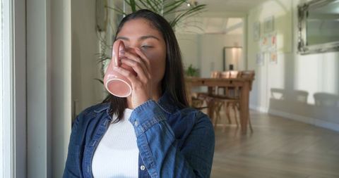 Asian woman sipping from pink mug by window in sunlit minimalist dining room with greenery