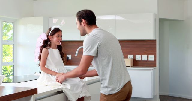 A man is playing with his young daughter in a modern kitchen. The daughter, dressed in a white dress and wearing fairy wings, sits on the counter while they share a joyful moment. This image can be used for concepts related to family bonding, parental love, playful activities at home, and modern family life.